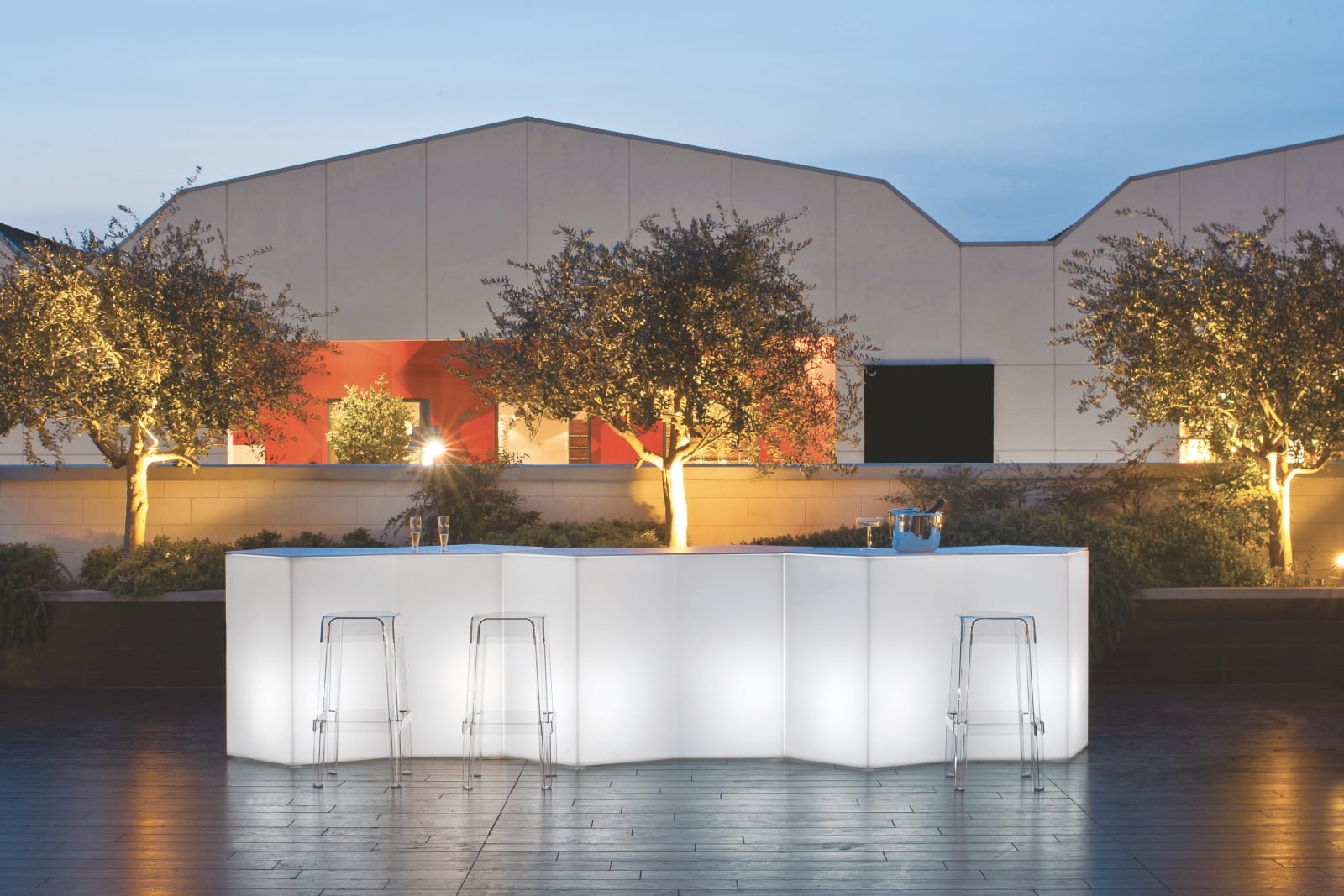 Modulare Leuchttheke Rubik Iceberg in Weiß für den Outdoor-Bereich mit transparenten Barhockern bei Dämmerung.